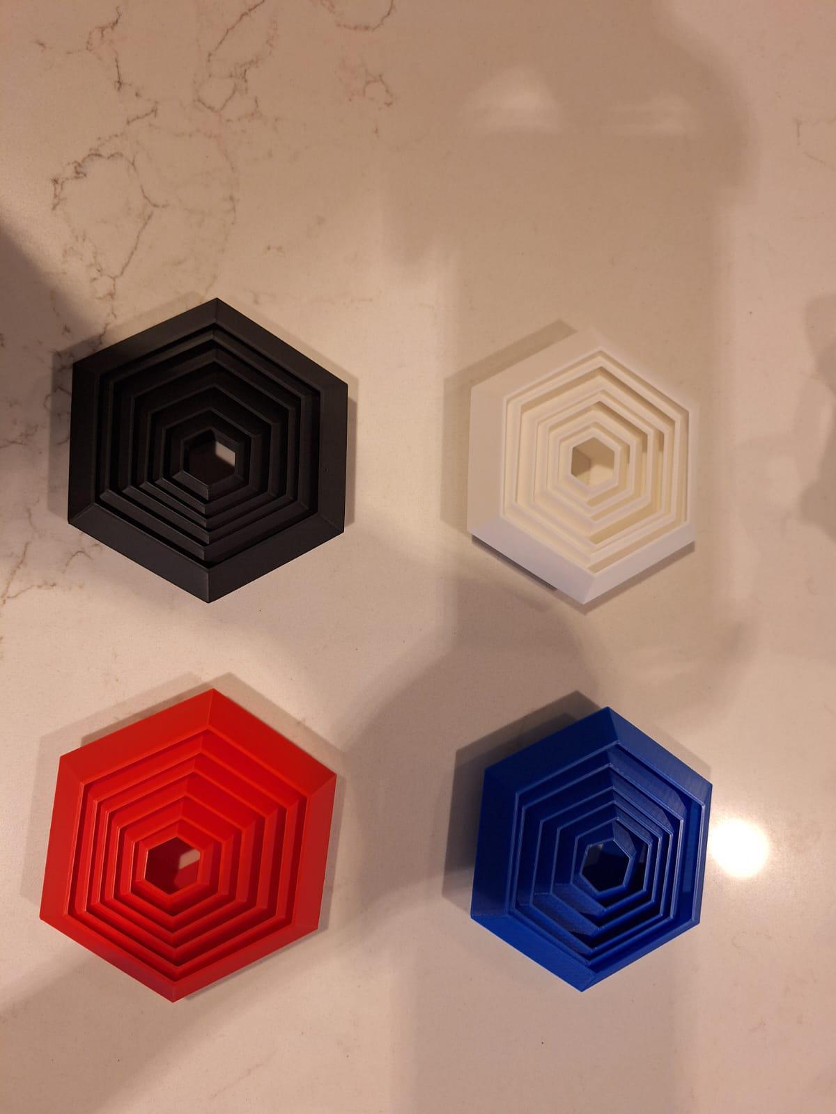 Set of four hexagonal 3D printed board game organizers in black, white, red, and blue on a marble surface