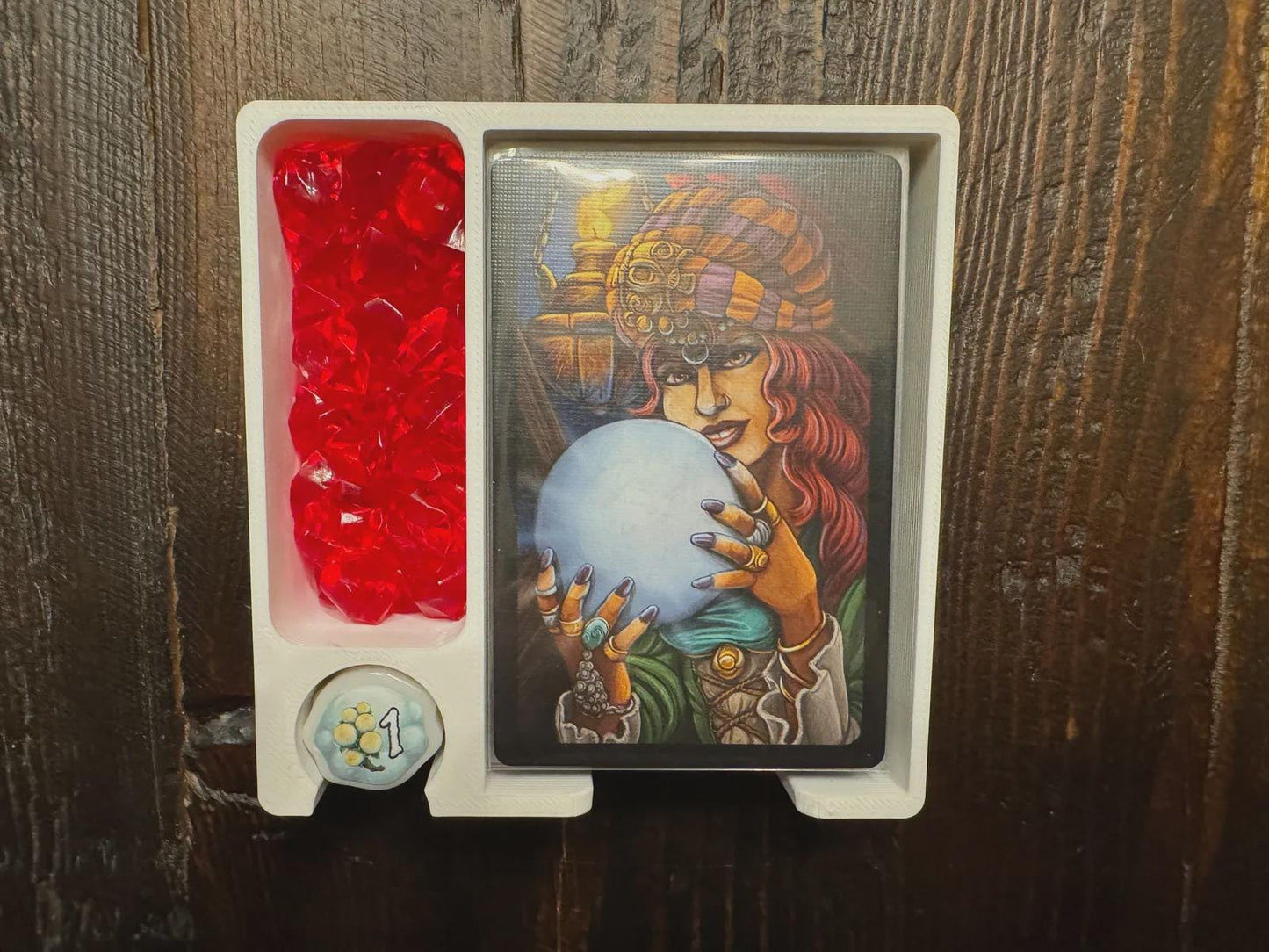 Board game organizer tray holding cards, red tokens, and a die, on a wood surface