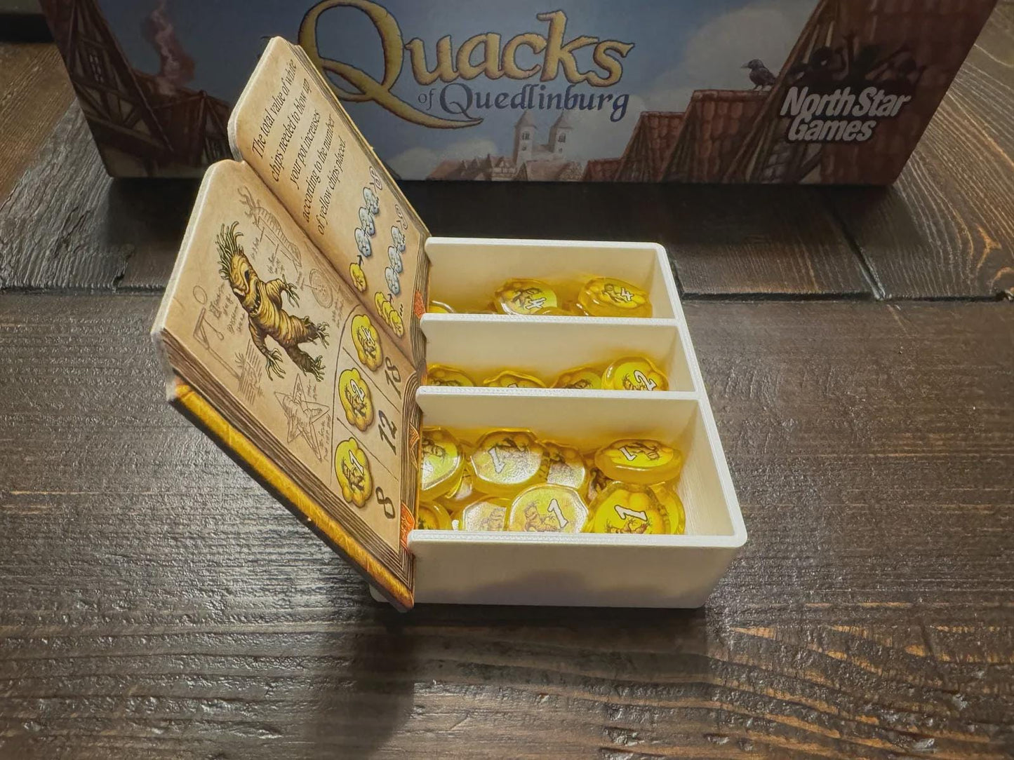 3D printed board game organizer for Quacks of Quedlinburg with gold tokens and open rulebook.