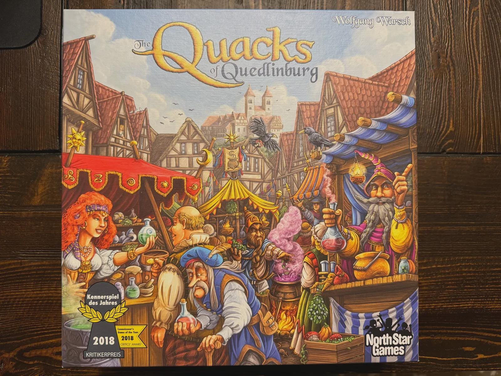 The Quacks of Quedlinburg board game box cover with colorful medieval market scene, displayed on wooden table. Board game storage and organizer shop KreativLayers.